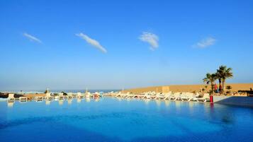 Une piscine d’un bleu éclatant, bordée de chaises longues et entourée de palmiers, avec une plage de sable fin et un ciel dégagé.