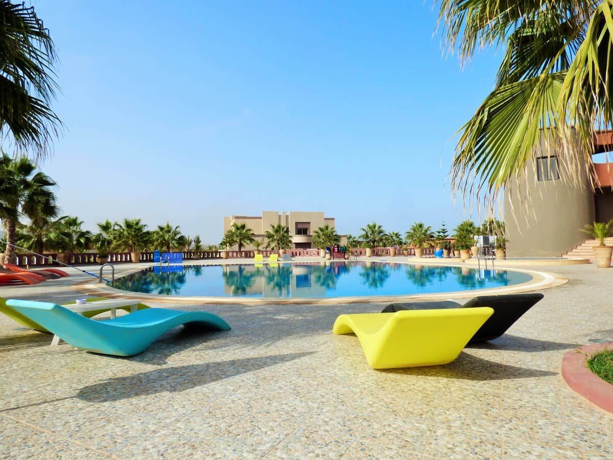 Una piscina con sillas de descanso, palmeras y un edificio al fondo.