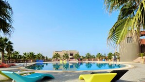 Piscina com espreguiçadeiras, palmeiras e um prédio ao fundo.