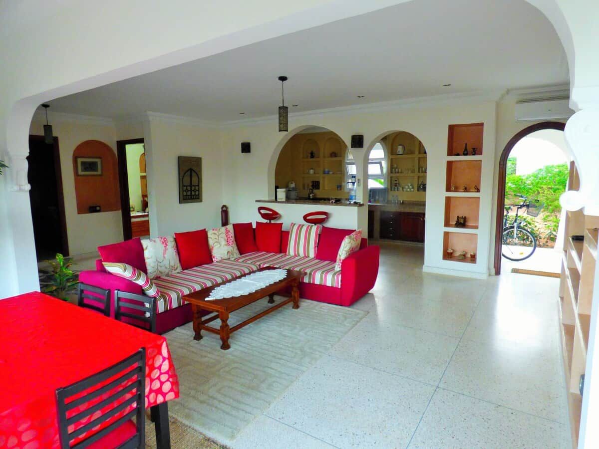A spacious living area with a red sofa set, a dining table, and a kitchen in the background.