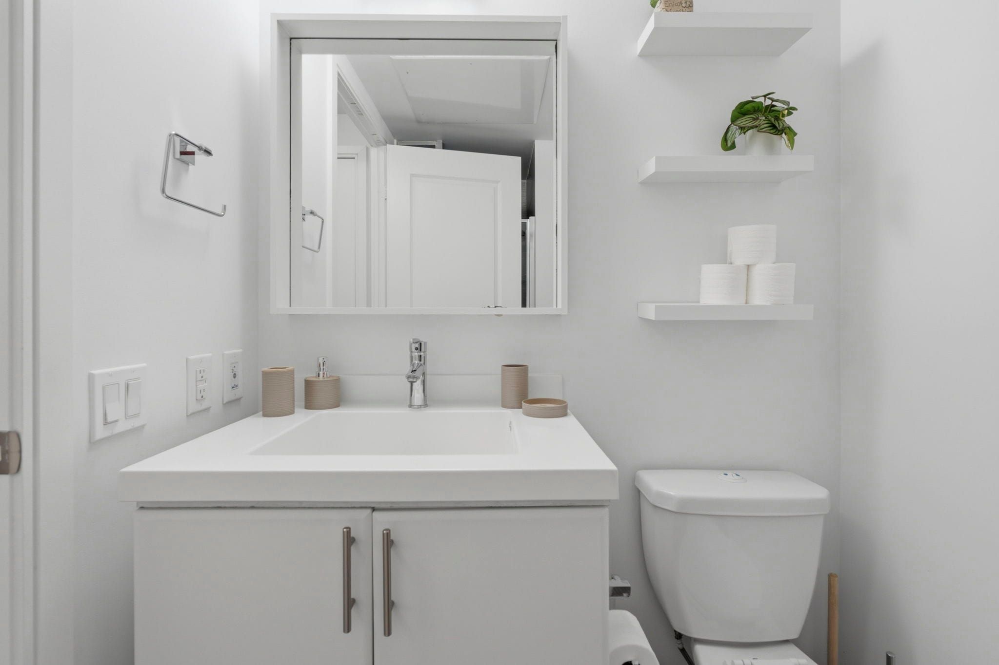 Un baño moderno con un lavamanos, espejo y retrete blancos. Hay estantes con artículos de aseo y una planta.