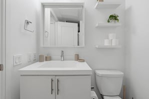 Un baño moderno con un lavamanos, espejo y retrete blancos. Hay estantes con artículos de aseo y una planta.