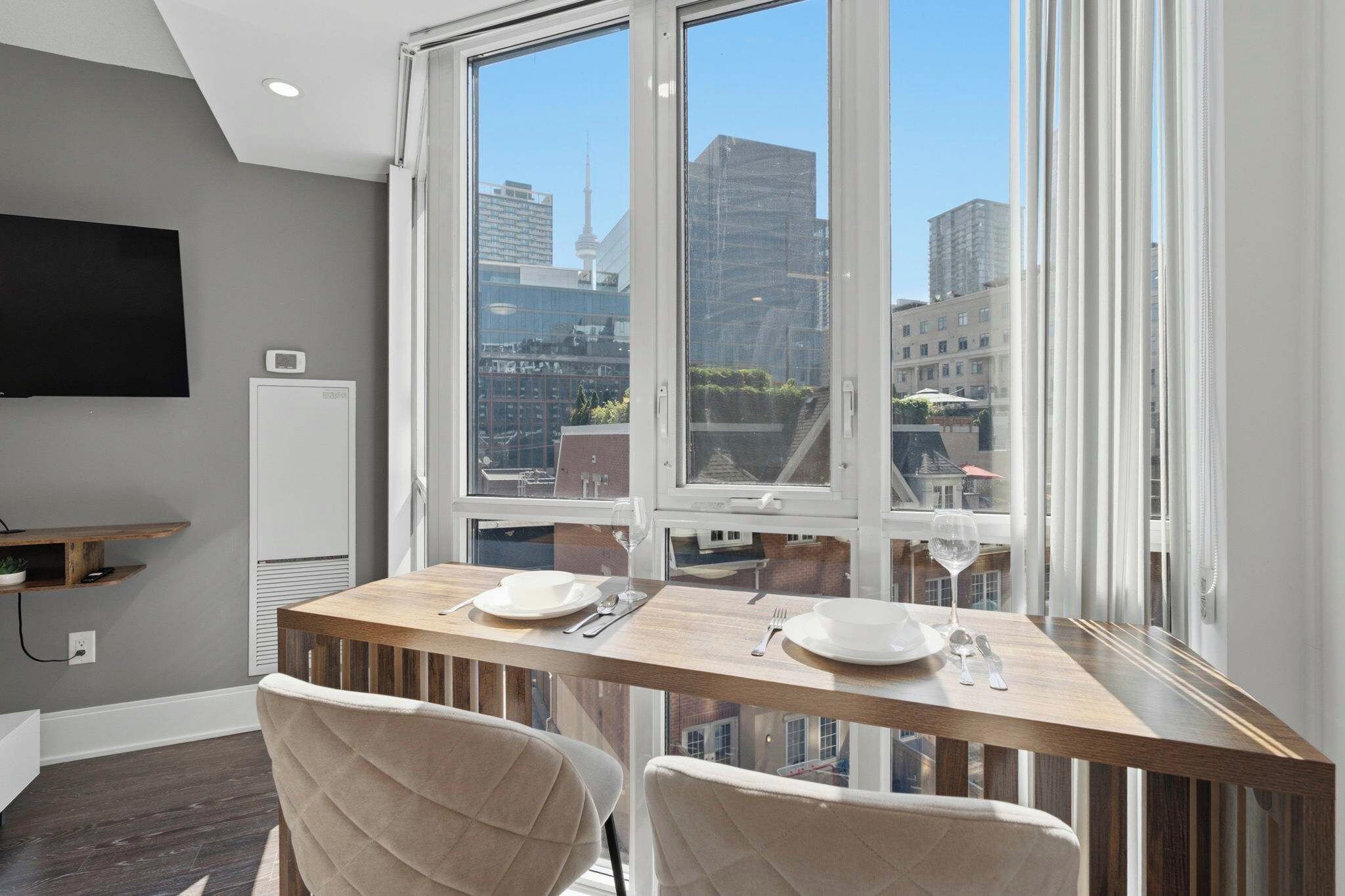 Un área de comedor con una mesa dispuesta para dos personas, un televisor en la pared y vistas panorámicas de la ciudad a través de amplios ventanales.
