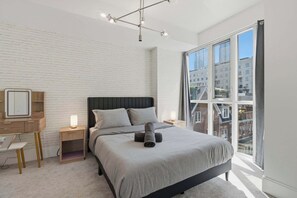 A modern bedroom with a large window, a bed with grey bedding, a wooden nightstand, a desk, and a mirror. - Modern 1BR - Walk to CN Tower - Sleeps 4 (Toronto)