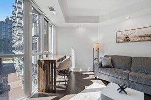 A modern living room with a sofa, a dining table, and a view of a cityscape through large windows. - Modern 1BR - Walk to CN Tower - Sleeps 4 (Toronto)