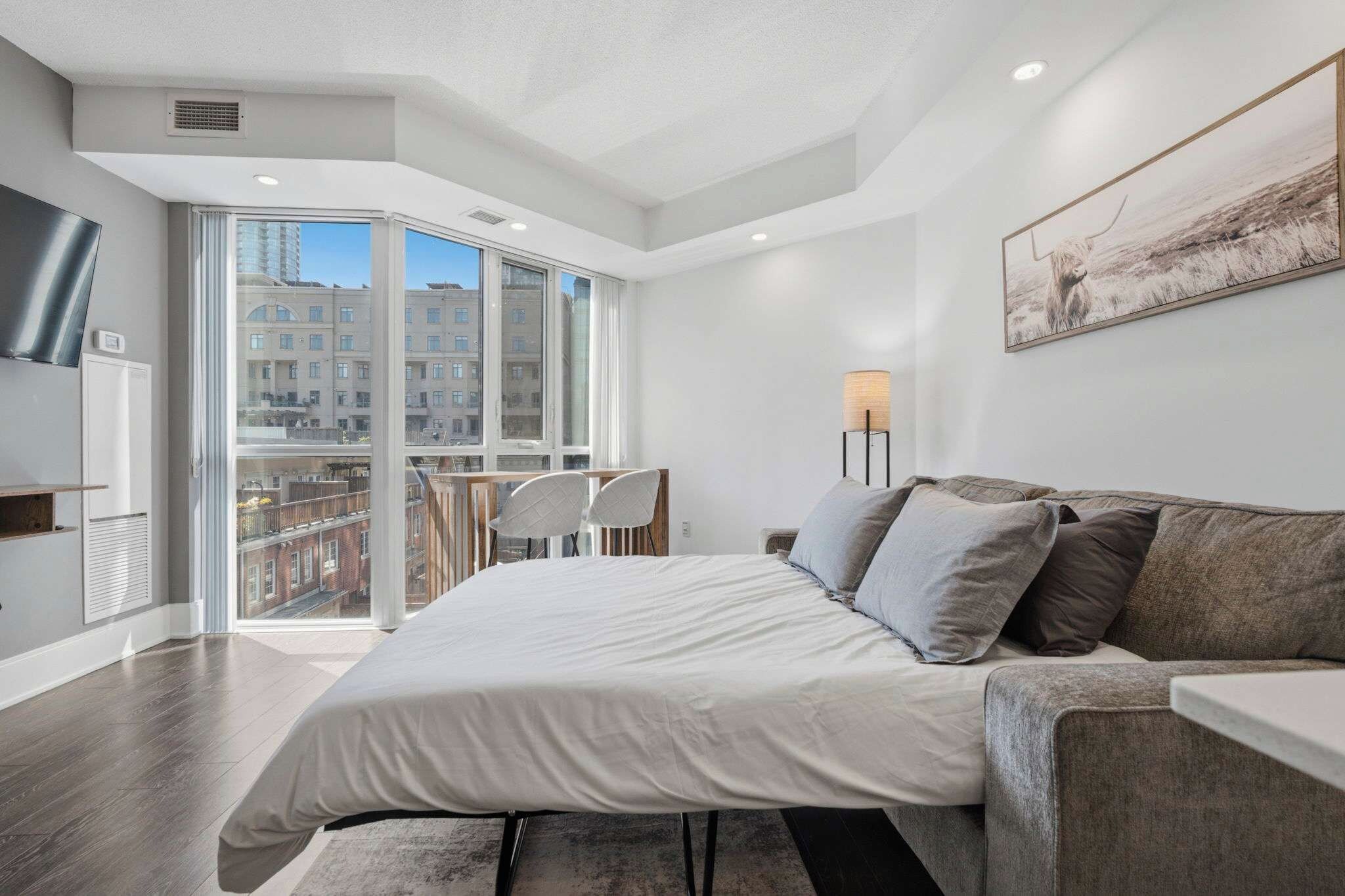 Un dormitorio moderno con una cama grande, un sofá, un escritorio y una silla. Se ve una ventana con vistas a edificios.
