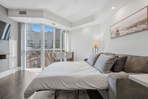 Un dormitorio moderno con una cama grande, un sofá, un escritorio y una silla. Se ve una ventana con vistas a edificios.