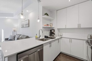 A modern kitchen with white cabinetry, a built-in dishwasher, a coffee machine, and a hanging light fixture. - Modern 1BR - Walk to CN Tower - Sleeps 4 (Toronto)