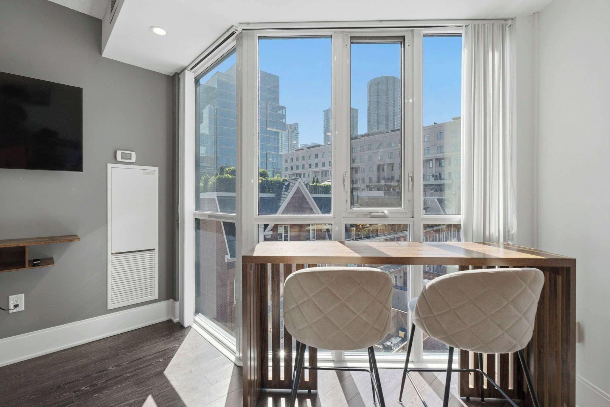 A modern room with a large window, a flat-screen TV, a table with two chairs, and a built-in heater.