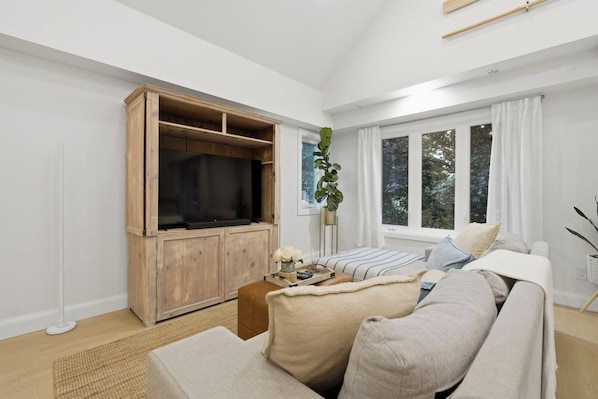 A modern living room with a wooden entertainment unit, a large window with curtains, a sofa, and a plant.