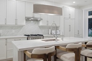 A modern kitchen with white cabinetry, a central island with bar stools, a stainless steel range hood, and a window providing natural light.