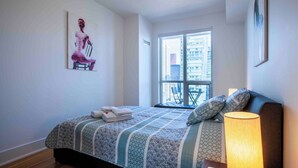 Una habitación de hotel con una cama, un ventanal con vistas a edificios, un cuadro en la pared y una lámpara en la mesita de noche.