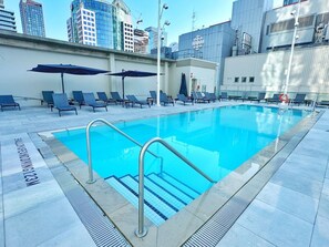 Ein Schwimmbecken mit Liegestühlen und Sonnenschirmen, umgeben von Stadtgebäuden.