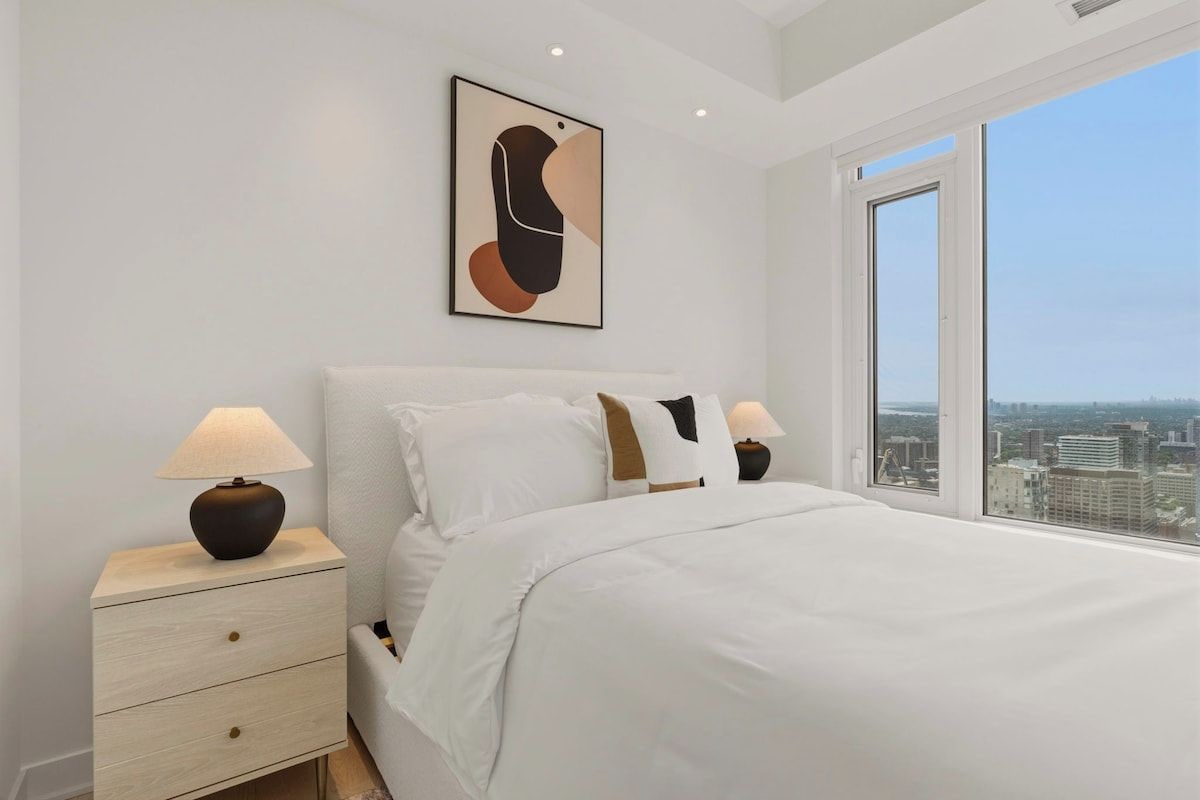 Un dormitorio con una cama grande, dos mesitas de noche con lámparas, vistas a la ciudad a través de un ventanal amplio y una obra de arte enmarcada en la pared.