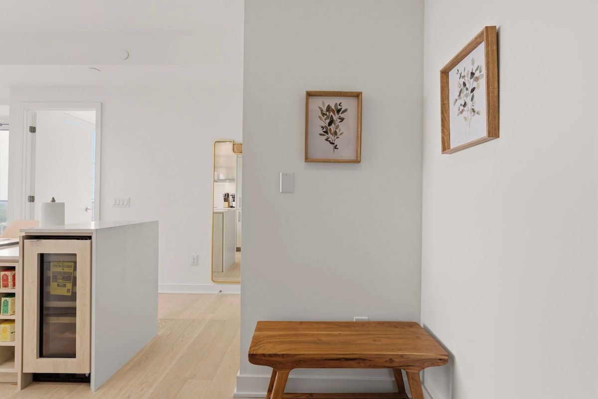 A modern interior with a wooden table, a white wall with framed artwork, and a kitchen area in the background.