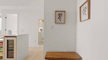 A modern interior with a wooden table, a white wall with framed artwork, and a kitchen area in the background.