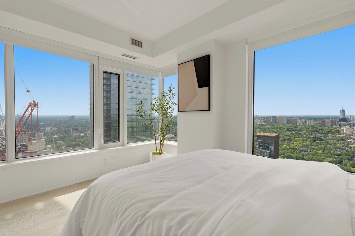 Un dormitorio moderno con una cama grande, una planta en maceta y vistas a la ciudad a través de amplios ventanales.