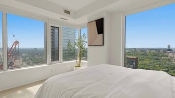 Un dormitorio moderno con una cama grande, una planta en maceta y vistas a la ciudad a través de amplios ventanales.