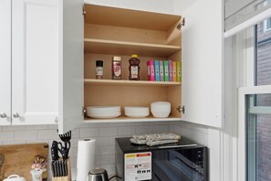 A kitchen with white cabinets, a wooden shelf with spices, a microwave, and a cutting board.