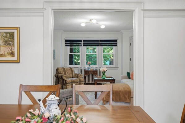 A dining area with a wooden table, chairs, and a vase of flowers. There is a window with blinds and a radiator in the background.