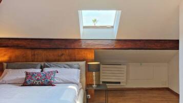 A bedroom with a skylight, a bed with pillows, a bedside table with a lamp, and a wall-mounted air conditioning unit.