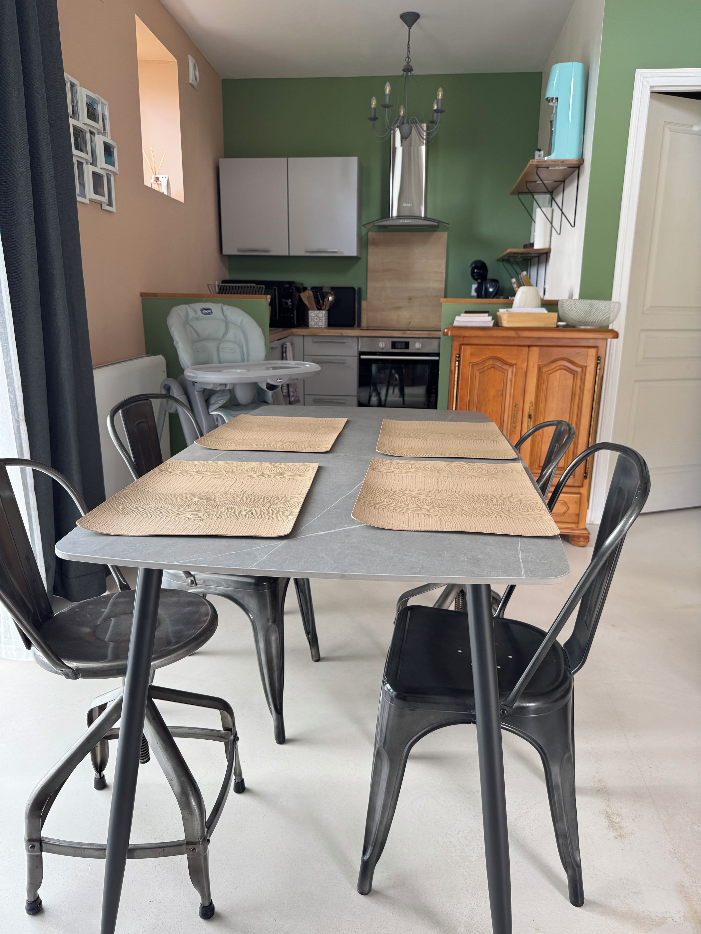 Un coin repas avec une table dressée pour quatre personnes, entourée de chaises, dans une pièce comprenant un coin cuisine et un mur d’accent de couleur verte.