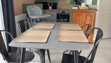 Un coin repas avec une table dressée pour quatre personnes, entourée de chaises, dans une pièce comprenant un coin cuisine et un mur d’accent de couleur verte.