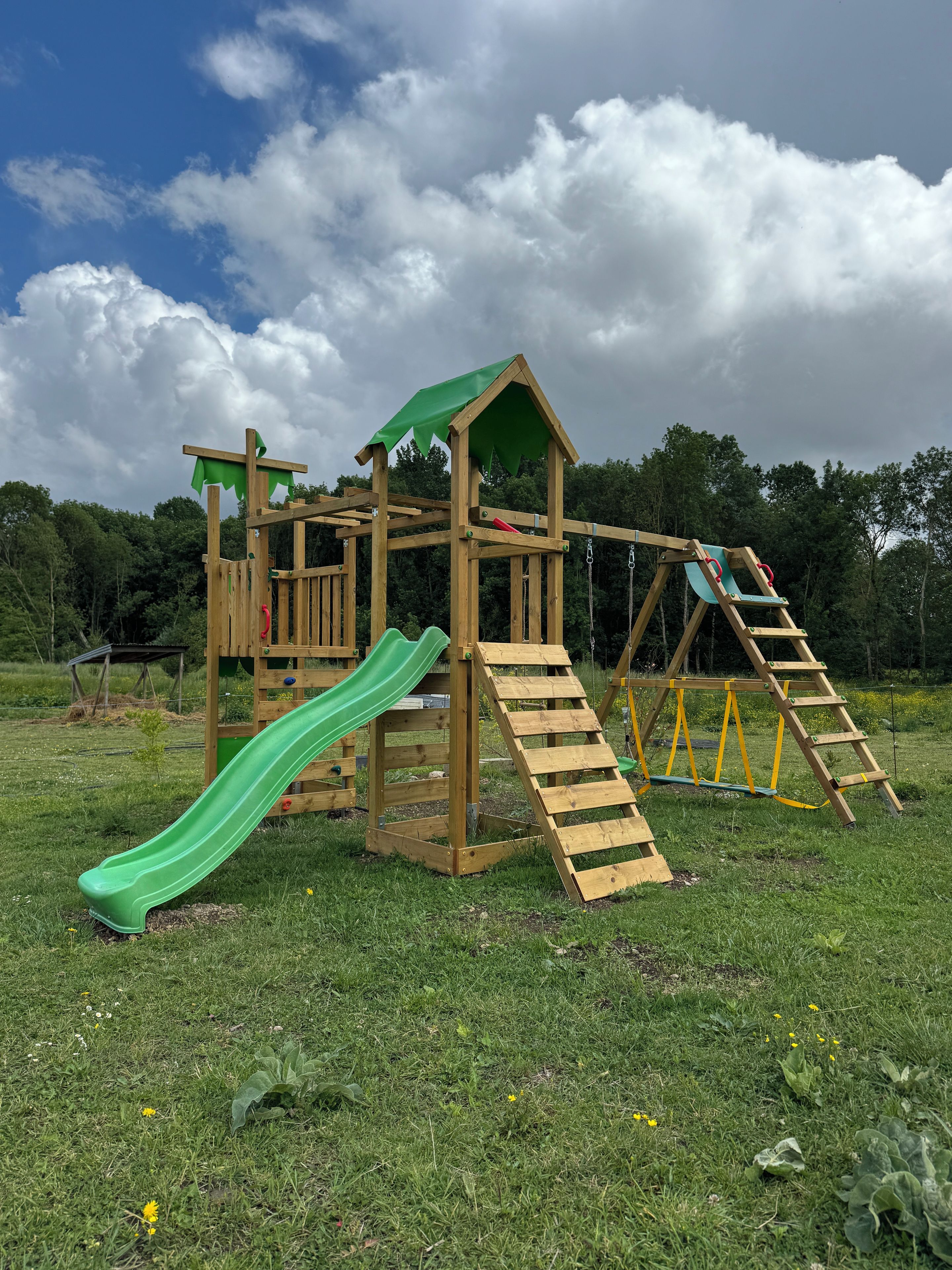Une structure de jeux en bois comprenant un toboggan, un filet d’escalade et un petit abri en forme de maison.