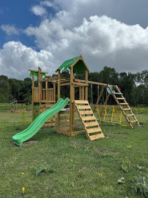 Une structure de jeux en bois comprenant un toboggan, un filet d’escalade et un petit abri en forme de maison.