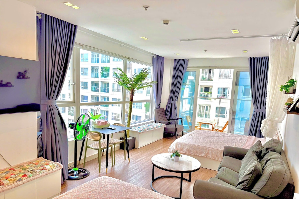 A modern hotel room with two beds, a dining table, a chair, a small round table, and a large window with curtains.
