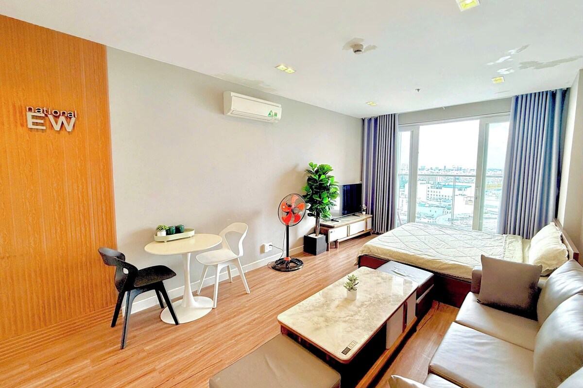 A hotel room with a bed, a sofa, a dining table, a TV, a plant, and a view of the city.