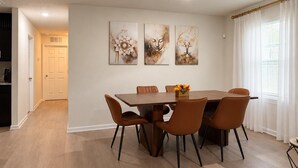 Een eetruimte met een houten tafel en zes stoelen, drie ingelijste kunstwerken aan de muur en een raam met gordijnen.