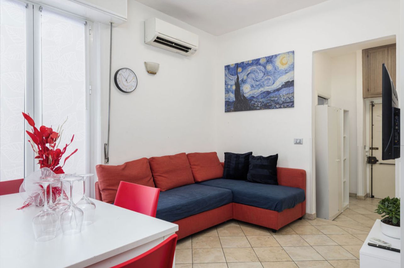 Un salon moderne avec un canapé d’angle rouge et bleu, une table à manger blanche avec des chaises rouges, une horloge murale et un tableau représentant un ciel nocturne.