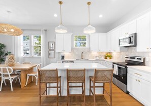 Une cuisine moderne avec des meubles blancs, un îlot central doté de tabourets de bar, un four à micro-ondes, une cuisinière et un luminaire suspendu.