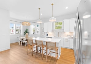 A modern kitchen with white cabinetry, a central island, and a dining area with wicker chairs. - The Bungalow: Private beach, Heated pool & hot tub, FREE Golf cart, Arcade Room (Miramar Beach)