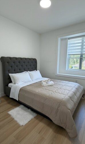 Une chambre à coucher avec un lit, des oreillers blancs, une tête de lit grise, une fenêtre avec des stores et un tapis blanc.