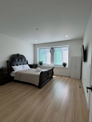 A bedroom with a bed, a nightstand, a window with blinds, a clock, and a TV mounted on the wall.