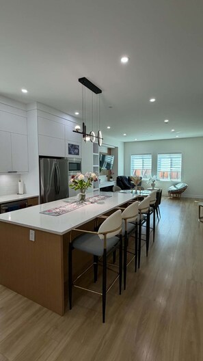 A modern kitchen with a central island, bar stools, a refrigerator, and a dining area with a table and chairs.