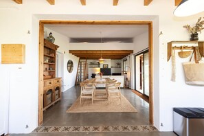 A spacious living area with a high ceiling, wooden beams, a dining table with chairs, a kitchen in the background, and a large window. - Ecofinca Lo Mas (Tortosa)