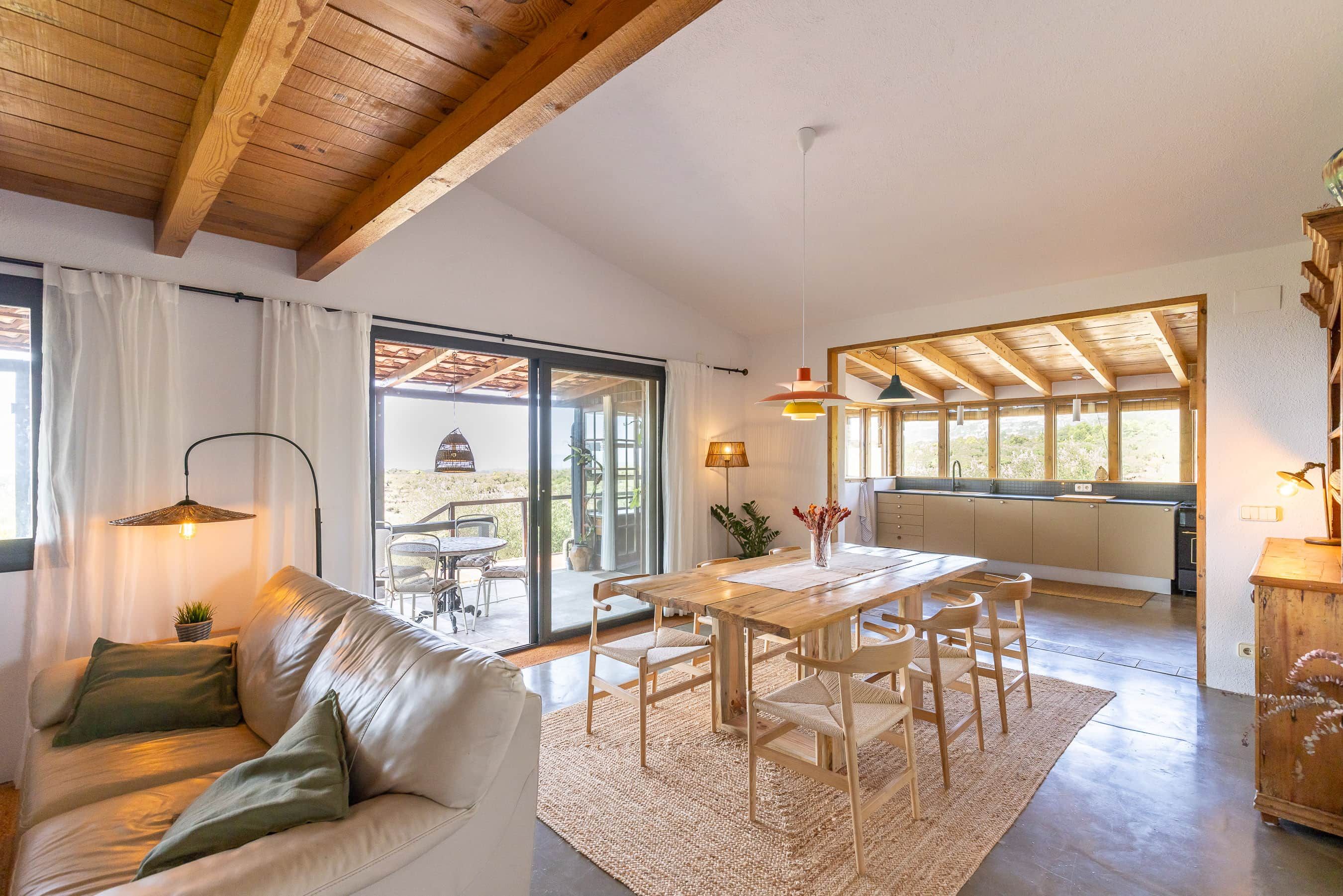 Amplia sala de estar con techo de madera, una mesa de comedor con sillas, un sofá con cojines y vistas a un patio exterior a través de un ventanal.