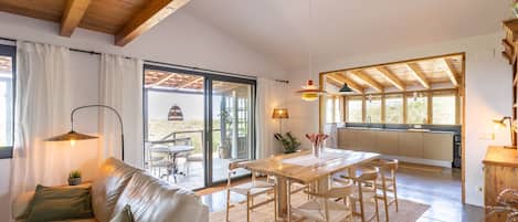 Amplia sala de estar con techo de madera, una mesa de comedor con sillas, un sofá con cojines y vistas a un patio exterior a través de un ventanal.