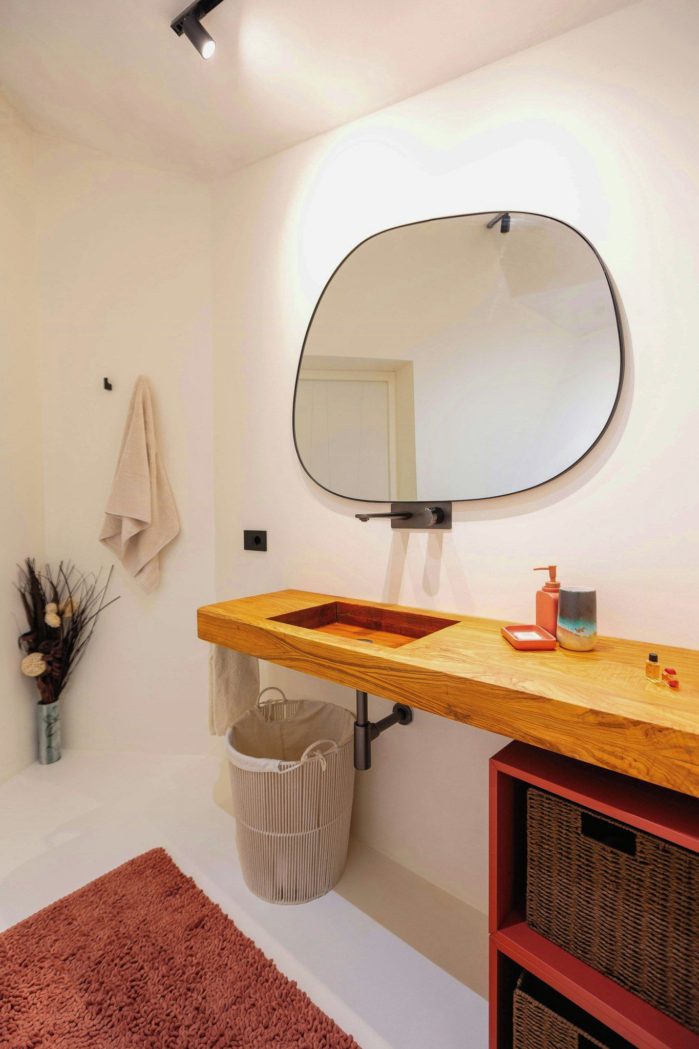 Un baño con tocador de madera, espejo redondo, toallero, cesto de basura y estante rojo.