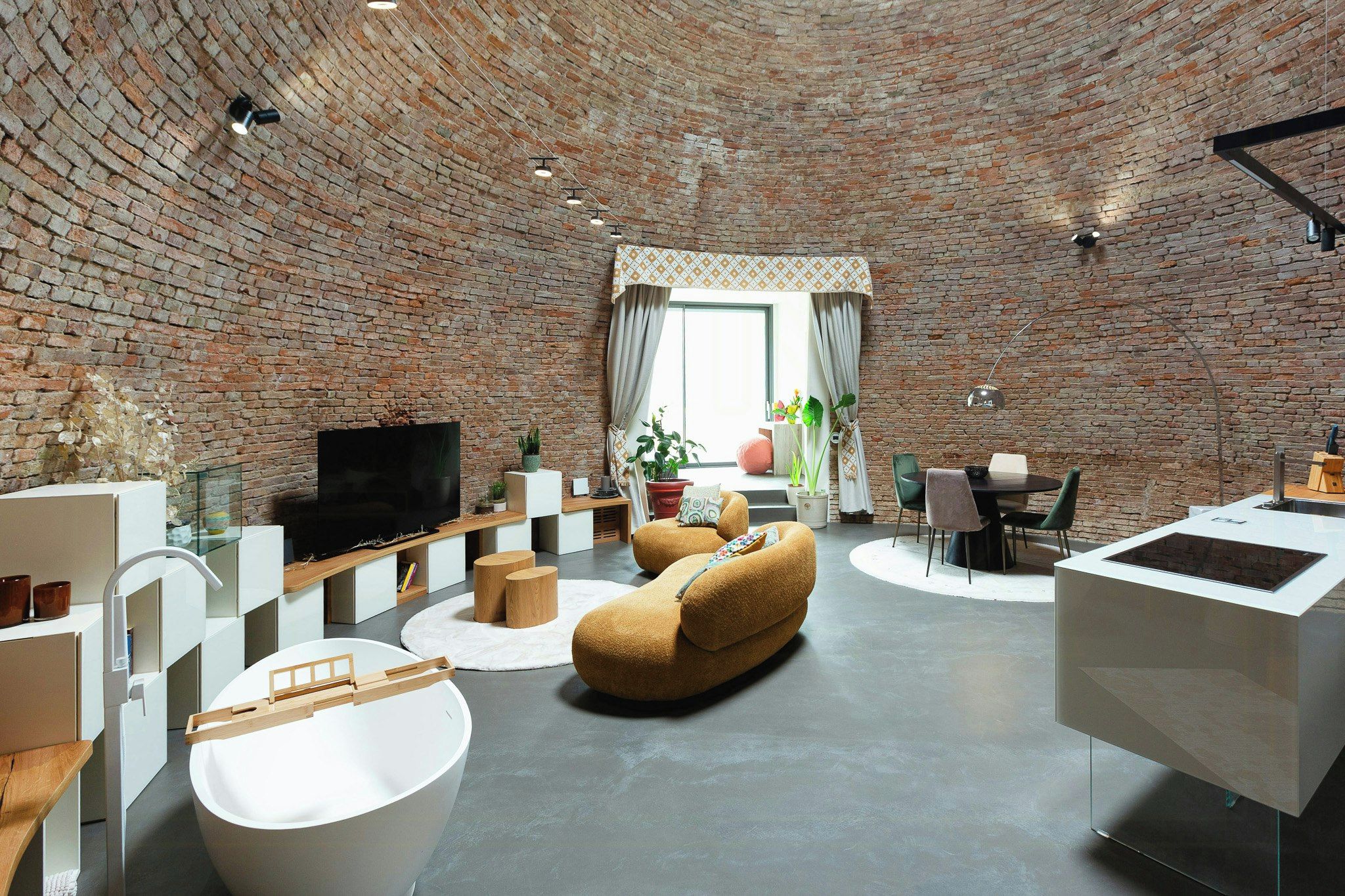 A modern living room with a brick wall, a flat-screen TV, a round coffee table, a sofa, and a dining area with chairs and a table.