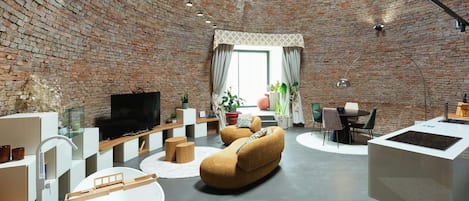 A modern living room with a brick wall, a flat-screen TV, a round coffee table, a sofa, and a dining area with chairs and a table.