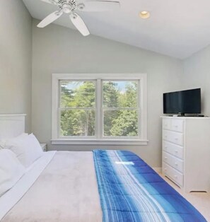 Un dormitorio con cama, televisor en una cómoda, ventana con vista a árboles y un ventilador de techo.