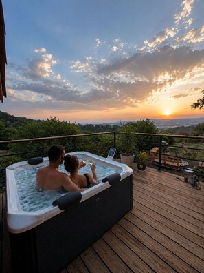 A hot tub on a wooden deck with a view of the sunset over a forested landscape.