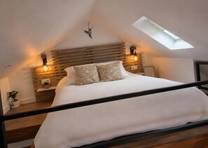 A bedroom with a sloped ceiling, a skylight, a bed with white bedding, wooden headboard, bedside lamps, and a small plant.