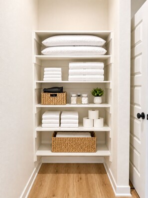 A white storage unit with shelves holding folded towels, a basket, and toiletries. - Goose Creek Modern Retreat (Farmington)