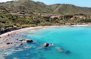 A coastal landscape with a beach, clear blue water, rocky shore, and a hillside with buildings.