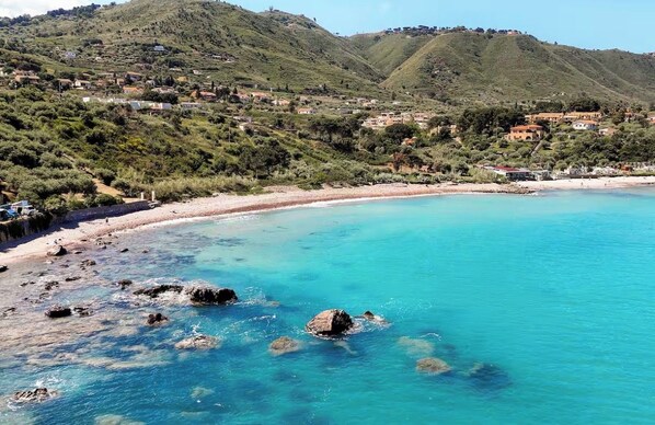 A coastal landscape with a beach, clear blue water, rocky shore, and a hillside with buildings.